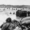 A view of Allied forces storming the beaches of Normandy from a ship used to transport soldiers in. Multiple soldiers are wading through the surf and some are climbing over the dunes to carry out the invasion.