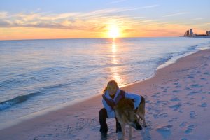The image shows the author crouching on a beach at sunset with her German shepherd Seeing Eye dog beside her. The dog is harnessed and the author has her left arm resting on the dog’s back. . The sky is filled with vibrant colors, transitioning from blue to orange as the sun sets behind the horizon. The sea is calm, with gentle waves lapping at the shore. The overall scene is peaceful and serene.