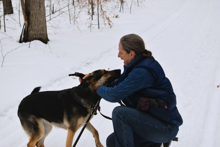 The author and her dog on a snow covered ski trail. She is crouching in the snow and gently holding the dog's face while she praises the dog.